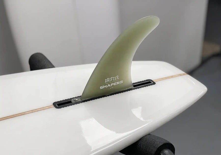 A close-up of a white surfboard's tail featuring a single, translucent green center fin secured in its fin box, showcasing a classic style of Longboard fins.