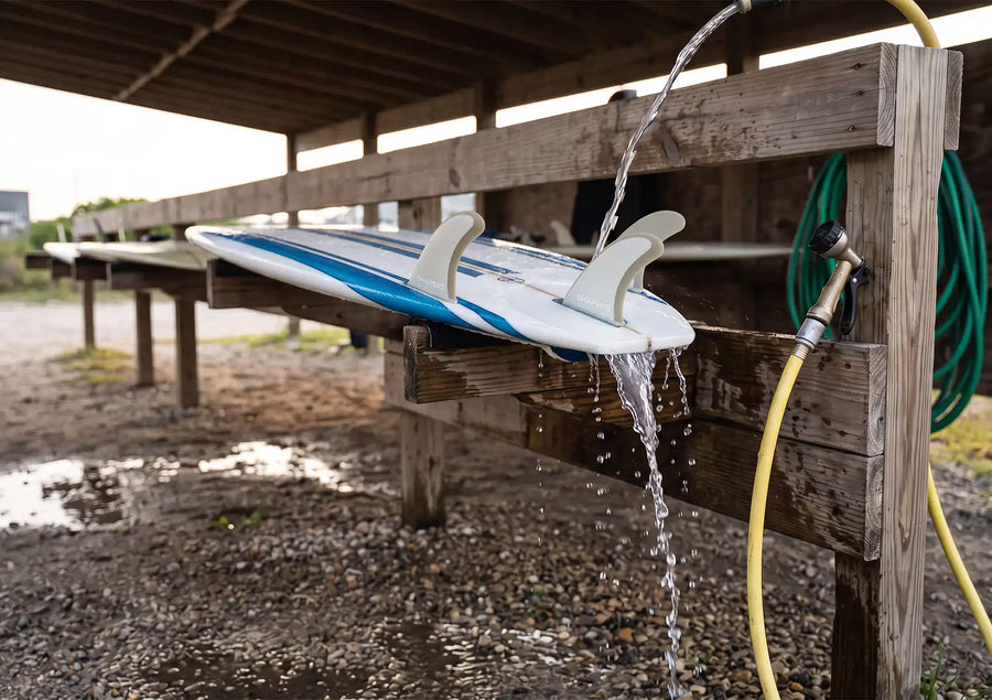 Alt text: Essential Surfboard Fin Care demonstrated by rinsing an upside-down blue and white surfboard and its attached fins with fresh water from a hose on a wooden outdoor rack.