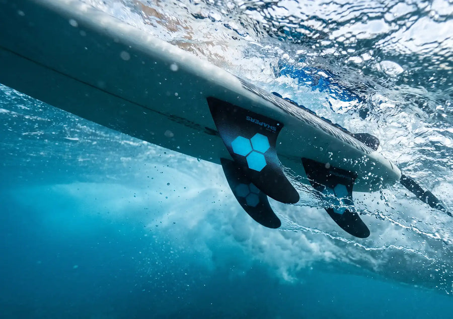 An underwater perspective looking up at the bottom of a surfboard as three black and blue Shapers fins slice through turbulent water, illustrating surfboard fins hydrodynamics in action on a wave.
