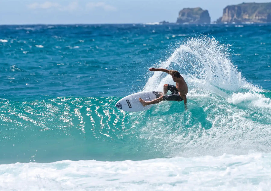 A surfer in black boardshorts executes a sharp, powerful turn on the lip of a bright blue wave, throwing a spray of white water and demonstrating the control provided by surfboard fins, with rocky islands visible in the background.