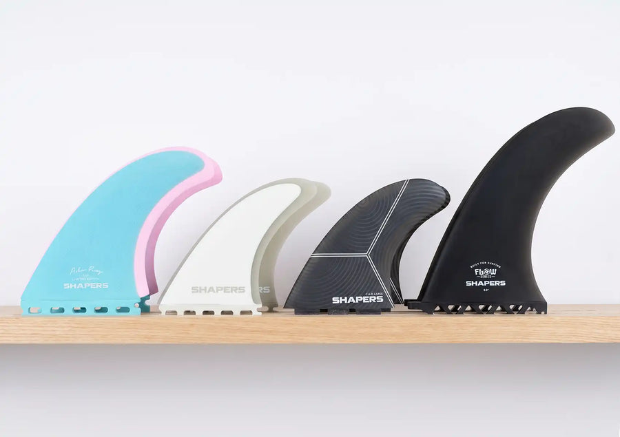 A visual comparison showing four different types of surfboard fins, ranging from colorful twin keels to a large black single fin, displayed on a wooden shelf against a white wall.
