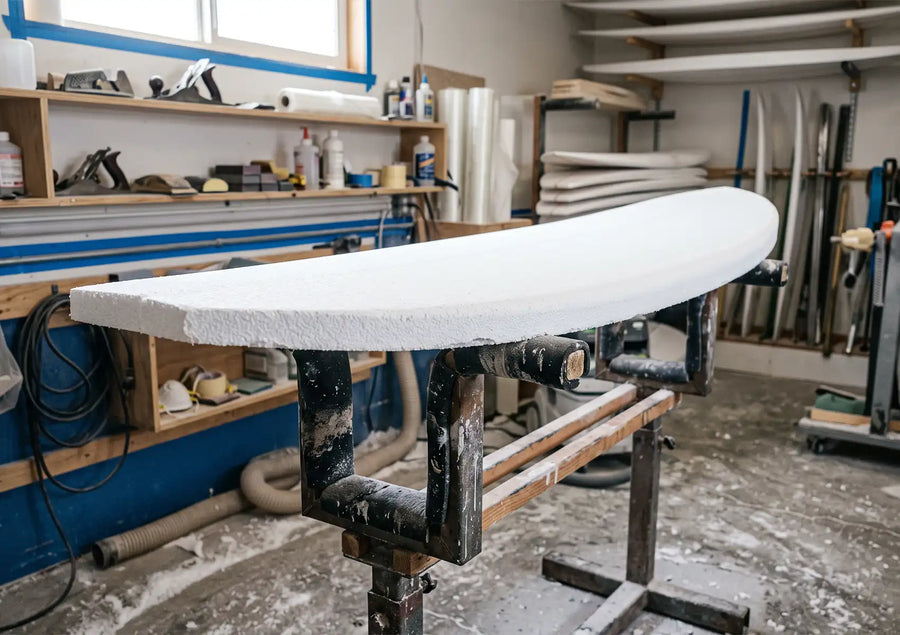 A solid white, unshaped surfboard blank resting horizontally on a padded shaping stand inside a workshop, with various tools, shelves, and foam blanks visible in the background.
