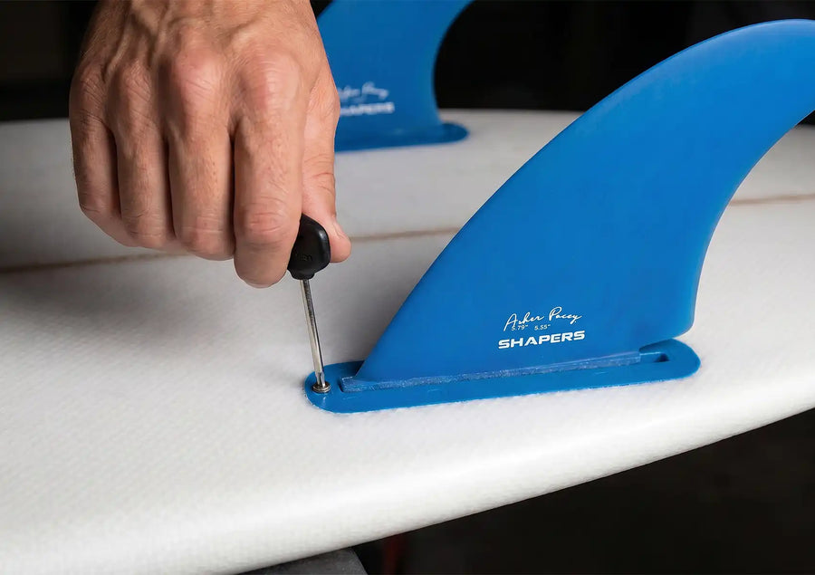 A close-up of a person using a black fin key to secure a blue Shapers fin into a surfboard, demonstrating the final step in how to install surfboard fins.